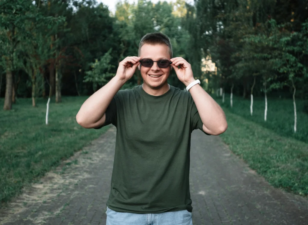 A cheerful young man wearing sunglasses and a green t-shirt, smiling on a pathway surrounded by trees.